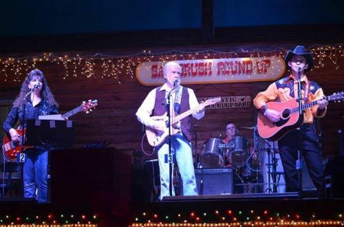 A country band performs on stage with guitars, drums, and festive lights in a rustic venue.