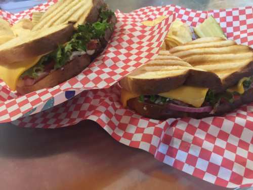Two grilled sandwiches with cheese, lettuce, and tomato, served with chips in red and white checkered baskets.