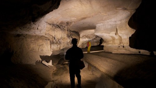 A person stands in a cave, silhouetted against the illuminated rock formations and water reflections.