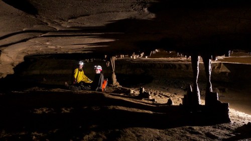 Two cavers sit on the ground in a dimly lit cave, surrounded by rock formations and water.