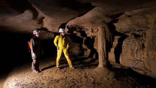 Two cavers in helmets explore a dimly lit cave with stalactites and stalagmites.