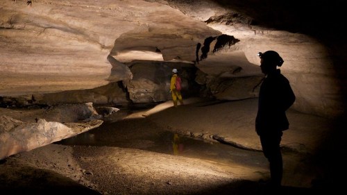 A person in a helmet stands in a dimly lit cave, with another figure in the background near a water pool.