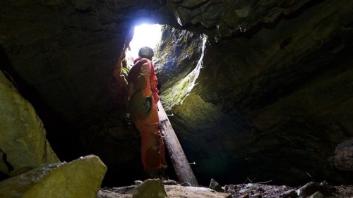 A person in an orange suit stands inside a cave, looking up at a bright opening above. Rocks and debris surround them.