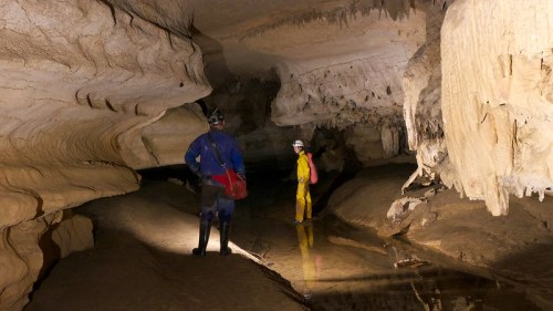 Two cavers explore a cavern with rock formations and a shallow water pool, illuminated by their headlamps.