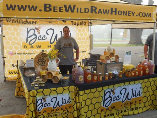 A vendor at a honey booth displays products, including jars of honey and honeycomb, under a yellow tent.