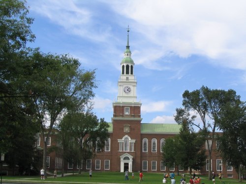 A historic brick building with a clock tower, surrounded by trees and people on a sunny day.