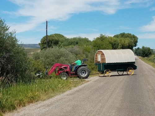 A red tractor pulls a green wagon along a rural road, surrounded by trees and grass under a blue sky.