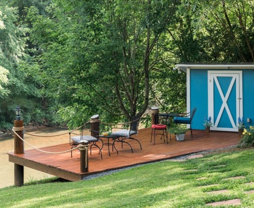 A cozy deck with chairs and a table beside a river, surrounded by lush greenery and a blue shed in the background.