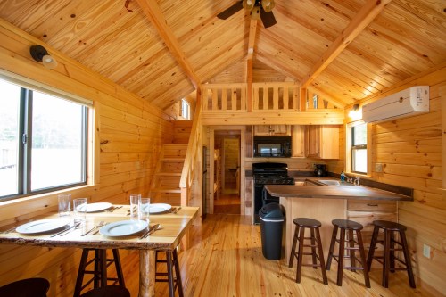 Cozy wooden cabin interior with a dining table, kitchen, and lofted sleeping area, featuring warm wood tones and natural light.
