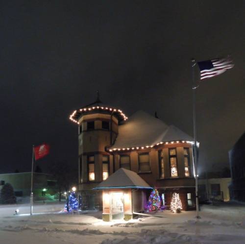 A snow-covered house with festive lights, surrounded by trees, under a night sky with flags waving.