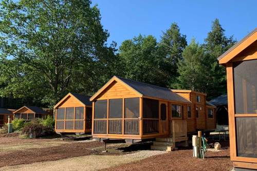 A row of wooden cabins with screened porches, surrounded by trees and a clear blue sky.