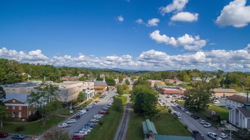 A scenic view of a small town with shops, trees, and mountains under a blue sky with fluffy clouds.