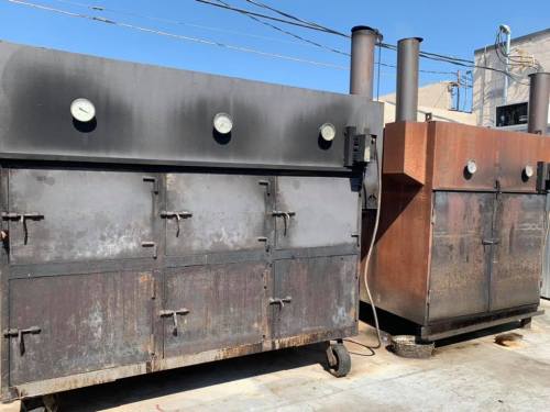 Two large, weathered smokers with gauges, positioned outdoors under a clear blue sky.