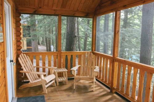 A cozy screened porch with two wooden chairs and a small table, surrounded by tall trees.