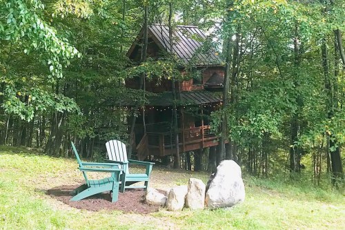 A cozy treehouse nestled in the woods, with a fire pit and green Adirondack chairs in the foreground.