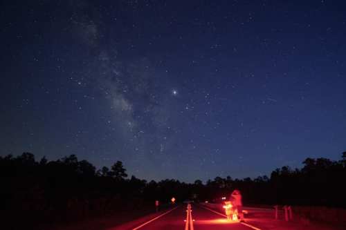 A dark road under a starry sky, with the Milky Way visible and a figure illuminated by red light.