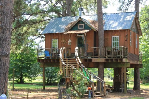 A charming treehouse with a wooden exterior, balcony, and slides, nestled among tall trees in a green landscape.