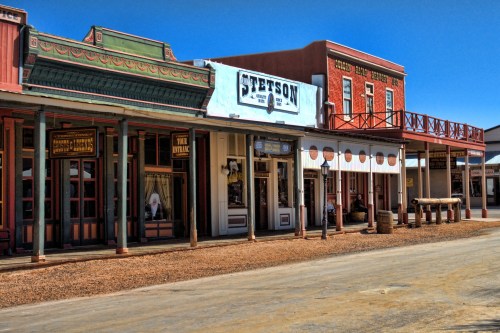A historic western-style street with colorful storefronts and a clear blue sky. Dusty road in the foreground.