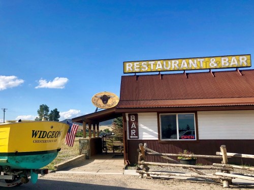 A rustic restaurant and bar with a yellow boat sign reading "Widgeon" and an American flag outside.