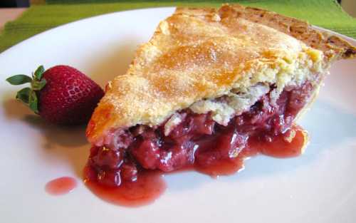 A slice of cherry pie with a flaky crust, oozing with cherry filling, served on a white plate with a strawberry.