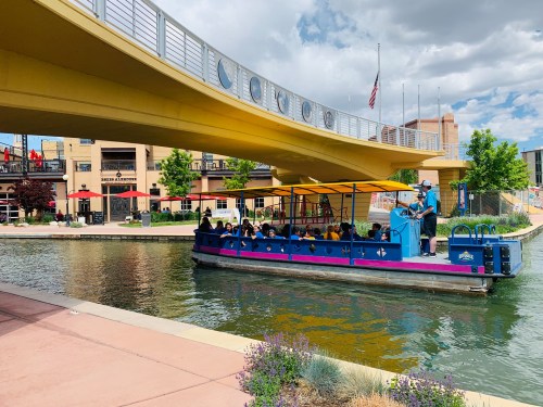 A colorful boat with passengers glides along a canal under a yellow bridge, surrounded by shops and greenery.
