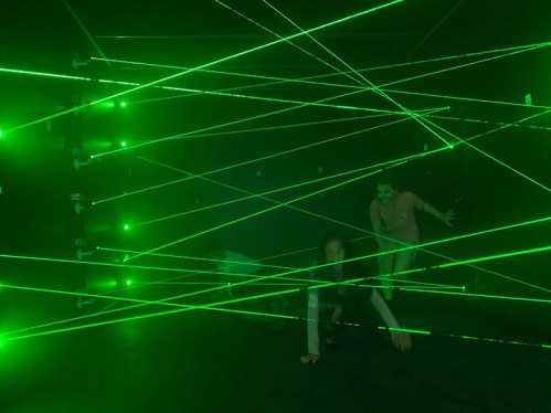 Two people navigate through a maze of green laser beams in a dark room.