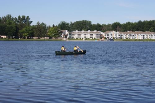 Two people paddle a canoe on a calm lake, with residential buildings visible in the background.