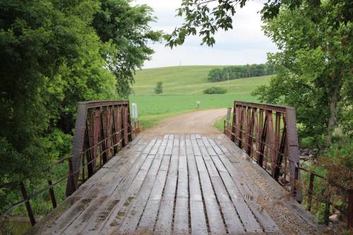 A rustic wooden bridge spans a dirt road, surrounded by lush greenery and rolling hills under a cloudy sky.