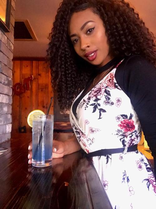 A woman with curly hair poses at a bar, holding a cocktail with a slice of lime, wearing a floral dress.