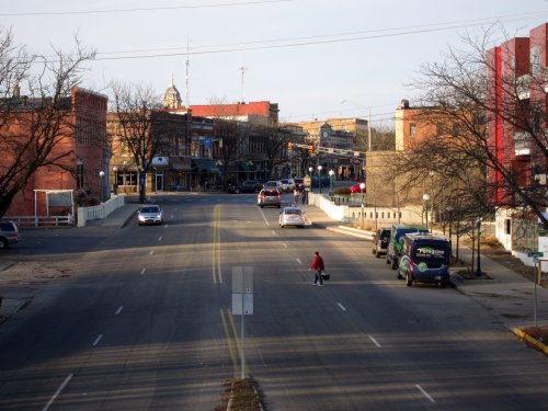 A quiet street scene with buildings on either side, a cyclist crossing, and cars parked along the road.