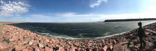 A panoramic view of a rocky shoreline with a calm sea under a blue sky, featuring a person standing on the rocks.