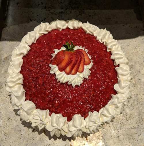 A round strawberry pie topped with whipped cream and fresh strawberry slices in the center.