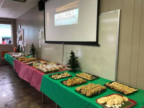 A table filled with various holiday cookies and treats, decorated with small Christmas trees, in a community room.