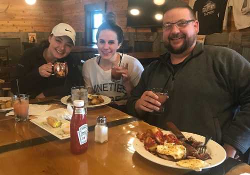 Three people smiling at a table with food and drinks, enjoying a meal in a cozy restaurant setting.