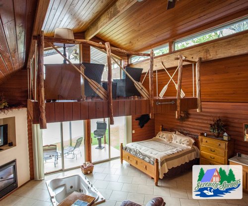 Cozy cabin interior with wooden beams, a loft area, and a hot tub, featuring large windows and a serene atmosphere.