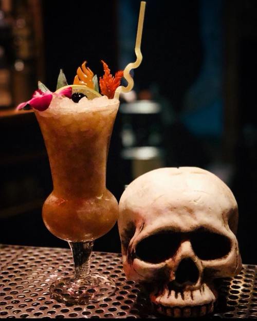 A colorful cocktail garnished with fruit and a straw, next to a decorative skull on a bar counter.