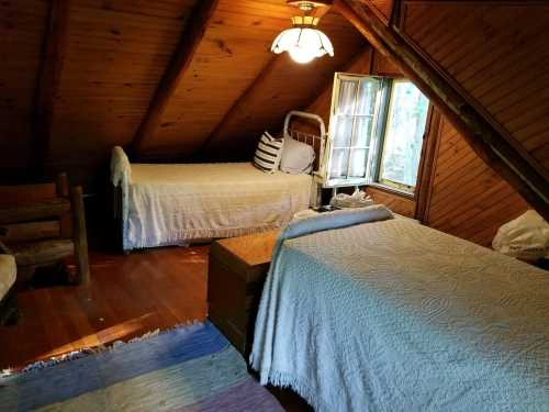 Cozy attic bedroom with two twin beds, wooden walls, a lamp, and a window letting in natural light.