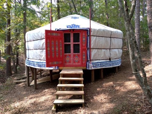 A traditional yurt with a red door and decorative trim, set in a wooded area with wooden steps leading up to it.