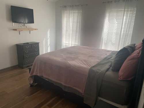 A cozy bedroom featuring a bed with pink and gray bedding, a dresser, and a wall-mounted TV. Natural light filters in through windows.