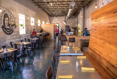 A spacious restaurant interior with wooden booths, metal tables, and diners enjoying their meals. Warm lighting enhances the ambiance.