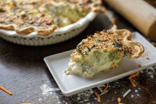 A slice of broccoli cheese pie on a plate, with the whole pie in the background and scattered cheese and flour around.