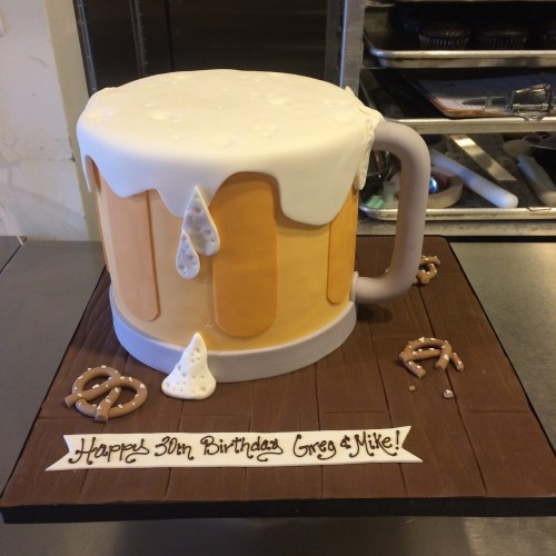 A cake shaped like a beer mug with frosting, pretzels, and a birthday message for Greg and Mike's 30th birthday.