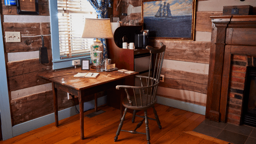A cozy room with a wooden desk, chair, lamp, and a painting, featuring rustic walls and warm wooden flooring.