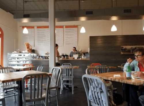 A cozy café interior with wooden tables, chairs, and a counter where staff prepare food. A customer enjoys a meal.