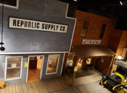 A vintage-style street scene featuring a supply store and a café, with wooden buildings and a classic car.