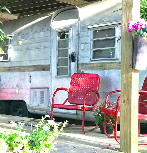 A vintage silver trailer parked under a wooden structure, with red chairs and potted flowers nearby.