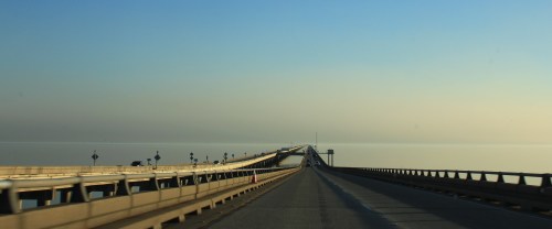 A long bridge stretches over calm water, leading into a hazy horizon under a clear blue sky.