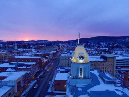 Aerial view of a snowy town at sunset, featuring a clock tower and illuminated buildings under a colorful sky.