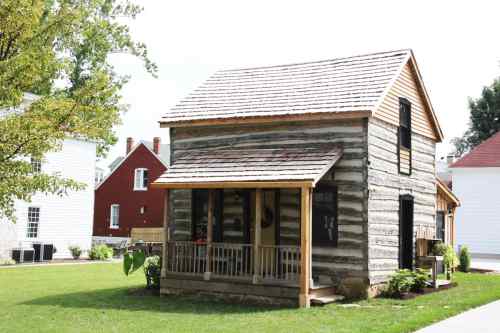 A small, rustic log cabin with a porch, surrounded by green grass and trees, set against a bright sky.