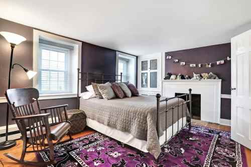 Cozy bedroom with a bed, rocking chair, and decorative fireplace, featuring purple walls and a patterned rug.